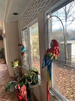 Wide shot of two ceramic parrots hanging on metal perches by window, showing overall size and color