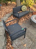 Full view of black woven wicker patio chair and matching bench with metal frames, with decorative round cushion on bench. Surfaces show leaves and signs of outdoor exposure.