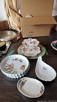 Several ceramic and porcelain plates on a table including Royal Worcester and Royal Winton pieces as well as a metal serving stand and a cardboard box in background.