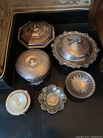 Six silver plated serving pieces arranged for display. Three covered serving bowls, two small decorative bowls, and one butter dish with glass insert.