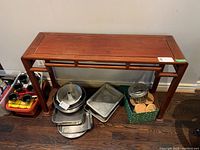 Front view of reddish-brown wood sofa table showing solid flat top and legs, with scuff marks visible on the wood surface. Various kitchen items including pans, cookie jars, trays, and coasters are placed beneath the table but not included.