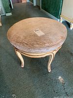 Full view of round foot stool showing beige upholstered textured top with wood base on carpet.