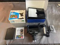 Overview photo showing one microscope with LCD screen, adjustable stand, USB cable, power adapter wrapped in plastic, manual, and open box.