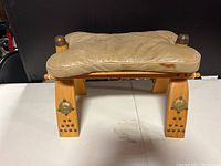 Front view of antique camel saddle showing leather cushion and wooden frame with metal studs and brass medallions.