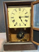 Open wooden clock case showing dial with Roman numerals and clock mechanism inside.