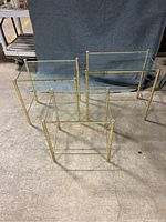 Three brass-framed nesting tables with glass tops arranged on concrete floor against a neutral backdrop. Shows the full set from front and above angles.