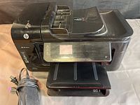 Front and side view of black HP Officejet 6500A Plus printer showing control panel, paper tray, and power cable.