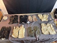 Eight pairs of gloves displayed on a grey surface with mix of black leather pilot and combat gloves, beige and light brown rawhide gloves in various sizes.