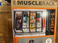 Front photo of the Musclerack storage unit in box showing the assembled unit with items on shelves and branding.