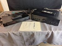 Photo showing Rotel stereo tuner BT-940AX, amplifier RB-980BX, two remote controls on top of amplifier, antenna wire, speaker wire, and manuals in protective sleeves on table with grey cloth and black backdrop.