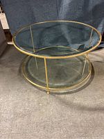 36 inch round two-tiered table with thick beveled glass tops and gold metal frame seen from an angled side view on concrete floor.