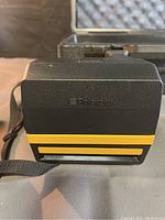Front view of the black and yellow Polaroid JobPro camera with wrist strap, placed in front of the foam-padded hard carrying case.