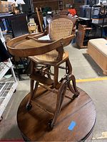 Side angle shot showing the decorative wooden arms, cane seat and back, spindled legs, wheels, and curved wooden tray attached to the highchair.