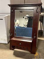 Front view of a wooden wall-mounted hall tree with a large mirror, metal hooks, and a small drawer beneath the mirror.