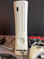 Front view of white Xbox 360 console standing vertically with power button and disc tray visible, with part of power cable and three controllers in background.