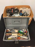 Photo of cardboard box filled with coiled power cords, cables, and electrical components. A grey tackle box filled with various hand tools is in front.