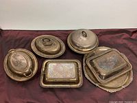 All five silver plated serveware items arranged on dark red fabric background, including lidded chafing dishes and tray.
