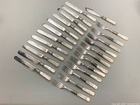 Top angle showing full set of 12 knives, 12 forks, and 1 fish cutting utensil with mother of pearl handles arranged neatly on gray surface.