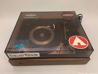 Photo showing turntable with closed dust cover featuring various stickers and power cord.