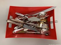 Top view photo of various vintage silver plated serving utensils placed in a red plastic basket. Items appear to include forks, knives, cake cutters and ladles with decorative handles.
