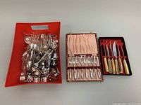Wide shot showing a large red plastic bin filled with assorted loose flatware, an 8-piece cake fork set in a pink satin-lined case, and a 6-piece steak cutting knife set with bone handles in a black and red tray.