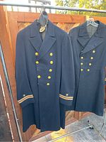 Photo showing two navy double-breasted military coats with gold buttons and single gold star insignia on sleeve cuffs hung on hangers outside