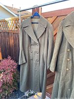 Photo showing one of the two heavy wool military coats hanging on a hanger outdoors, beige/olive colored with double-breasted buttons and wide lapels.