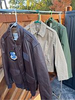 Three military jackets on hangers hung on metal rod outdoors.