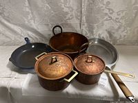 Five cookware items displayed: two small copper saucepans with lids, large copper stockpot, blue skillet, aluminum pan.