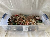 Clear plastic bin filled with numerous strands and pieces of costume jewelry including bracelets, necklaces, and bangles in a mix of colors and materials.