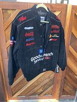 Black leather racing jacket hanging on a wooden fence, displaying front logo detail including Goodwrench Service Plus, Coca-Cola, Oreo, Snap-on, ACDelco, GM logos.