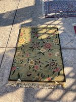 Green floral patterned woven rug with fringe edges, laid out on a cement floor.