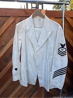 Front view of white US Navy dress uniform jacket showing double-breasted design and Chief Petty Officer rating badge on right sleeve.