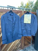 Two blue military style jackets hanging on hangers with visible front pockets, button closures, and collar.