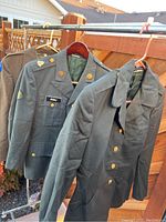 Three olive green military uniform jackets on hangers displayed on a rack outdoors, showing details including gold buttons and name patch 'Mccull'.