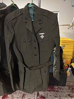 Front view of dark green military jacket with button closure, epaulets on shoulders, flap pockets, and medal insignia on chest pocket.