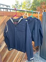 Three navy blue sailor shirts hanging on hangers outdoors against a fence. Each shirt has white stripes on collar and cuffs.