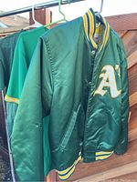 Front angle view of green Oakland Athletics satin varsity jacket with 'A' logo on chest, hanging on metal rod outside