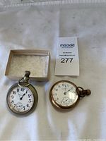 Photo of two pocket watches with white faces and metal cases on white background, one next to small box and auction tag