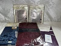 Photo of two Lillian brand lace panels in packaging, a Ralph Lauren tablecloth in dark burgundy with floral pattern, and two other packaged tablecloths, blue and purple.