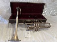 Full view of vintage silver-finish trumpet with attached mouthpiece resting on a white fabric surface alongside a black hard case with red velvet interior opened behind it.