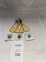 Photo shows three vintage milk tokens on white cards with handwritten text indicating producer, distributor, milk type, and volume. Above the tokens are five cream-colored milk bottle-shaped tags with printed and handwritten information, all arranged on a light fabric.