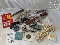 Wide photo showing watches, watch straps, vintage coins, pearl necklace, and accessories arranged on white cloth.