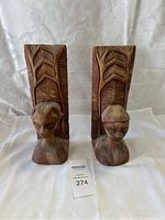 Front view of pair of wood carved bookends with figurative heads and geometric pattern backs placed side by side on white fabric background.