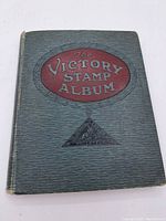 Front cover of The Victory Stamp Album, showing the title in a red oval on a dark textured background, with a decorative triangular emblem below.