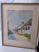 Framed watercolor painting showing a village street, houses, people, and chimney smoke under a light blue sky.