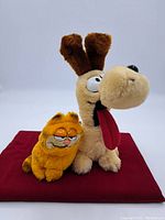 Both Garfield and Odie stuffed animals together on a red cloth, front facing