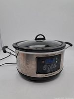 Front view of stainless steel Hamilton Beach slow cooker with glass lid and black side handles.