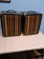 Pair of vintage 1970s portable speakers with brown patterned fabric covers and black casing with corner protectors and handles on top, shown side by side on a table.