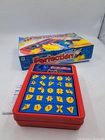 Full view of Perfection game with red plastic base, yellow shaped pegs in a blue grid, and original box behind it.